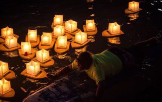 memorial service candles floating on water at night, with man on surfboard guiding them