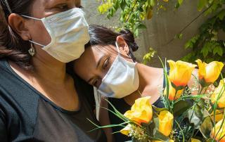 mother and daugher wearing covid masks and holding yellow flowers at outdoor funeral in cemetery in LA